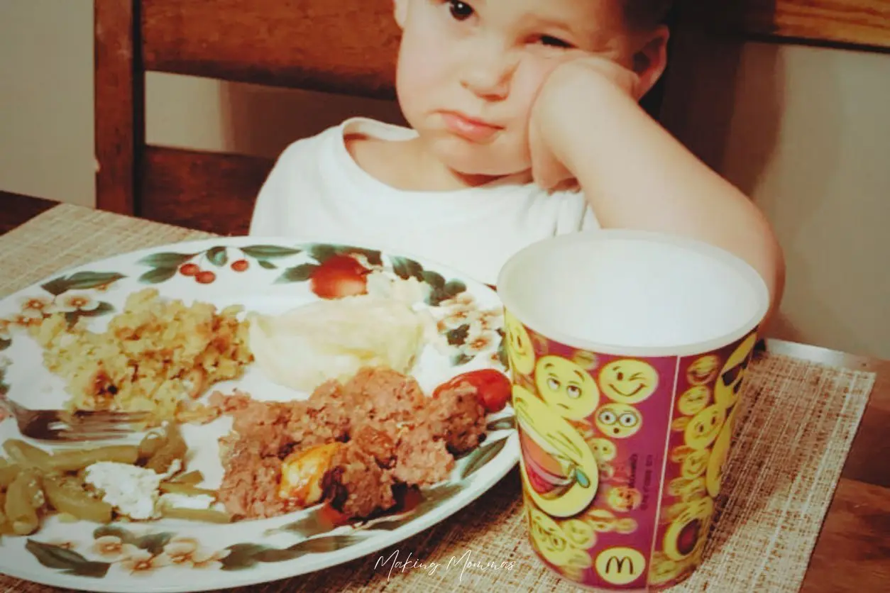 image of a disappointed looking small boy sitting at the table with a plate of food and a plastic cup with emoji smiley faces all over it