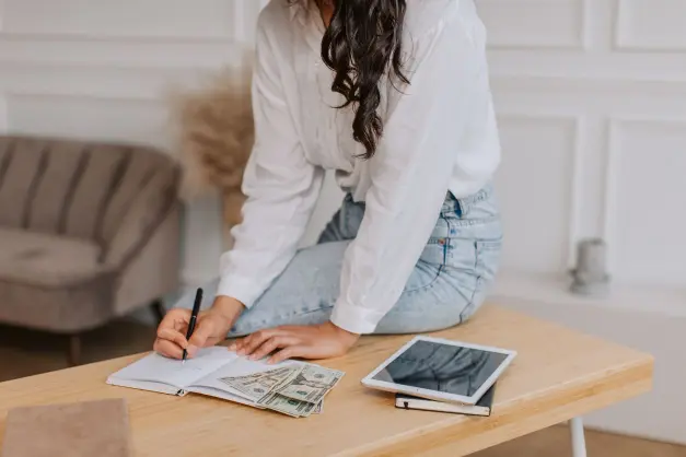 image of a woman with long hair sitting on a table, writing something in a notebook with money, an ipad, and a notebook next to her