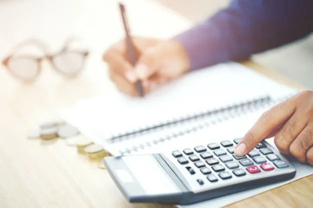 image of someone using a calculator, notebook, and pen with coins on the table