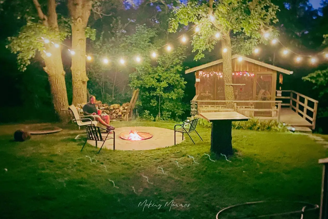 image of a man sitting at a firepit with a screened in porch in the background and lights strung through the trees at night