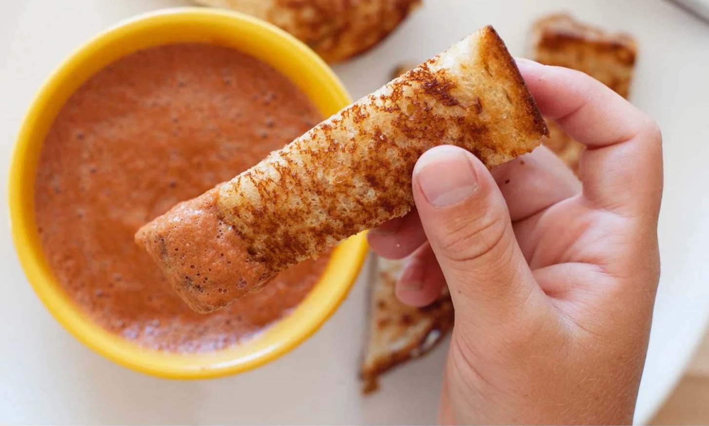 image of a hand dipping a grilled cheese dipper into a cup of tomato soup.