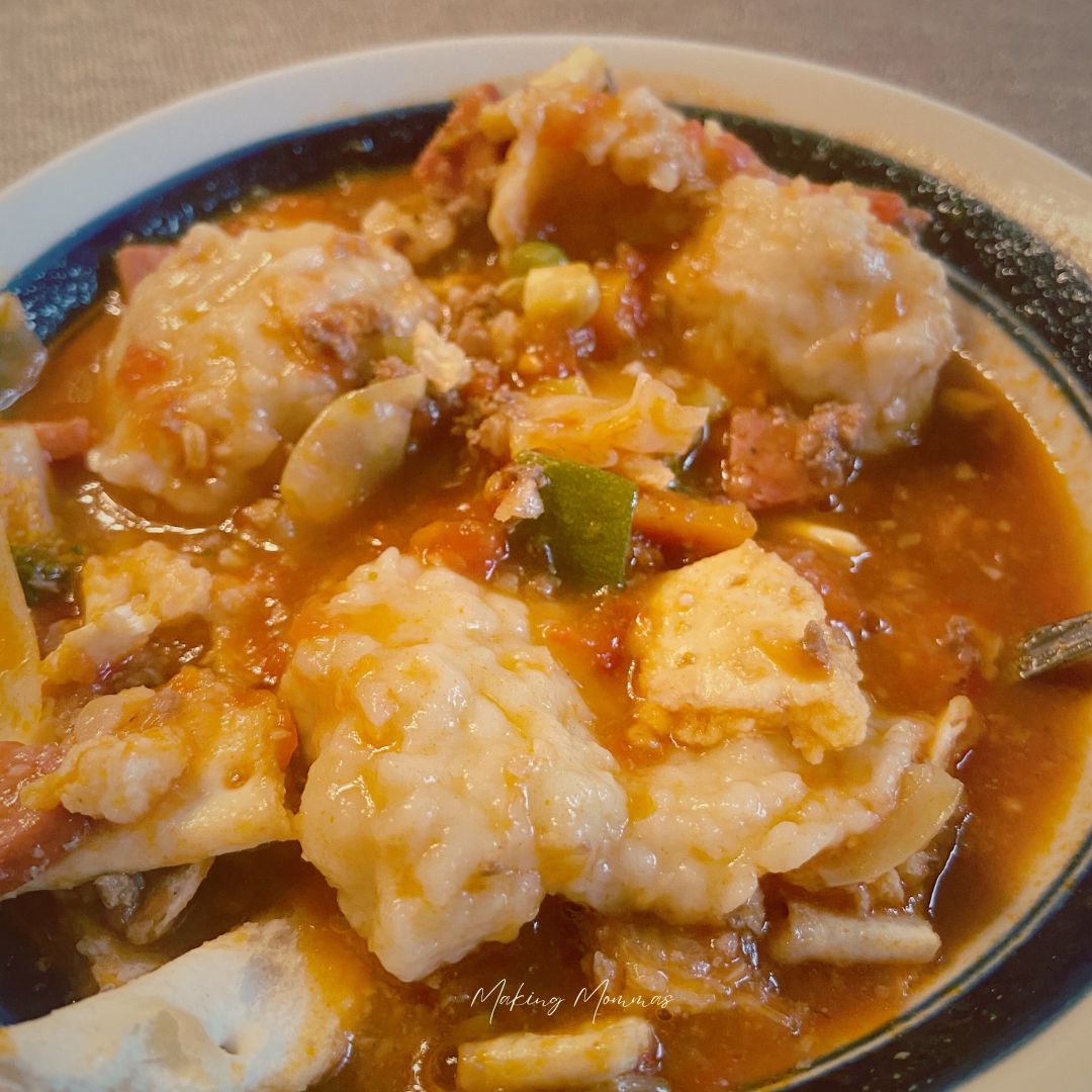 A hearty bowl of homemade ground beef vegetable soup, filled with colorful vegetables and topped with fluffy dumplings, served in a white bowl with a dark rim. The soup has a rich, tomato-based broth and is garnished with chunks of tender beef and dumplings, perfect for a cozy, comforting meal.