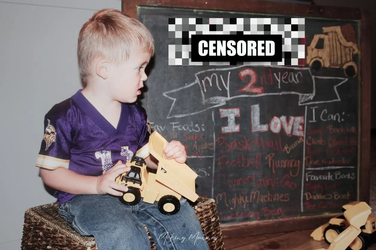 image of a little boy sitting on a basket next to a chalk board sign that has his age on it. His name is censored. He's holding a dump truck.