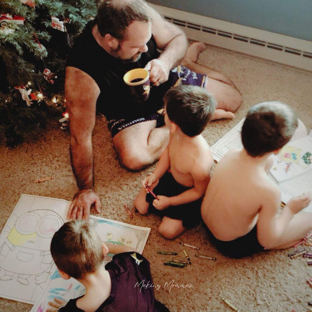 Image of three boys coloring on the living room floor with their dad, in front of the Christmas tree