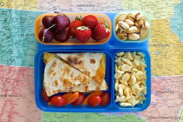 image of a quesadilla packed in a bento box with carrots and tomatoes, strawberries and cherries, nuts, and goldfish, laying on a map