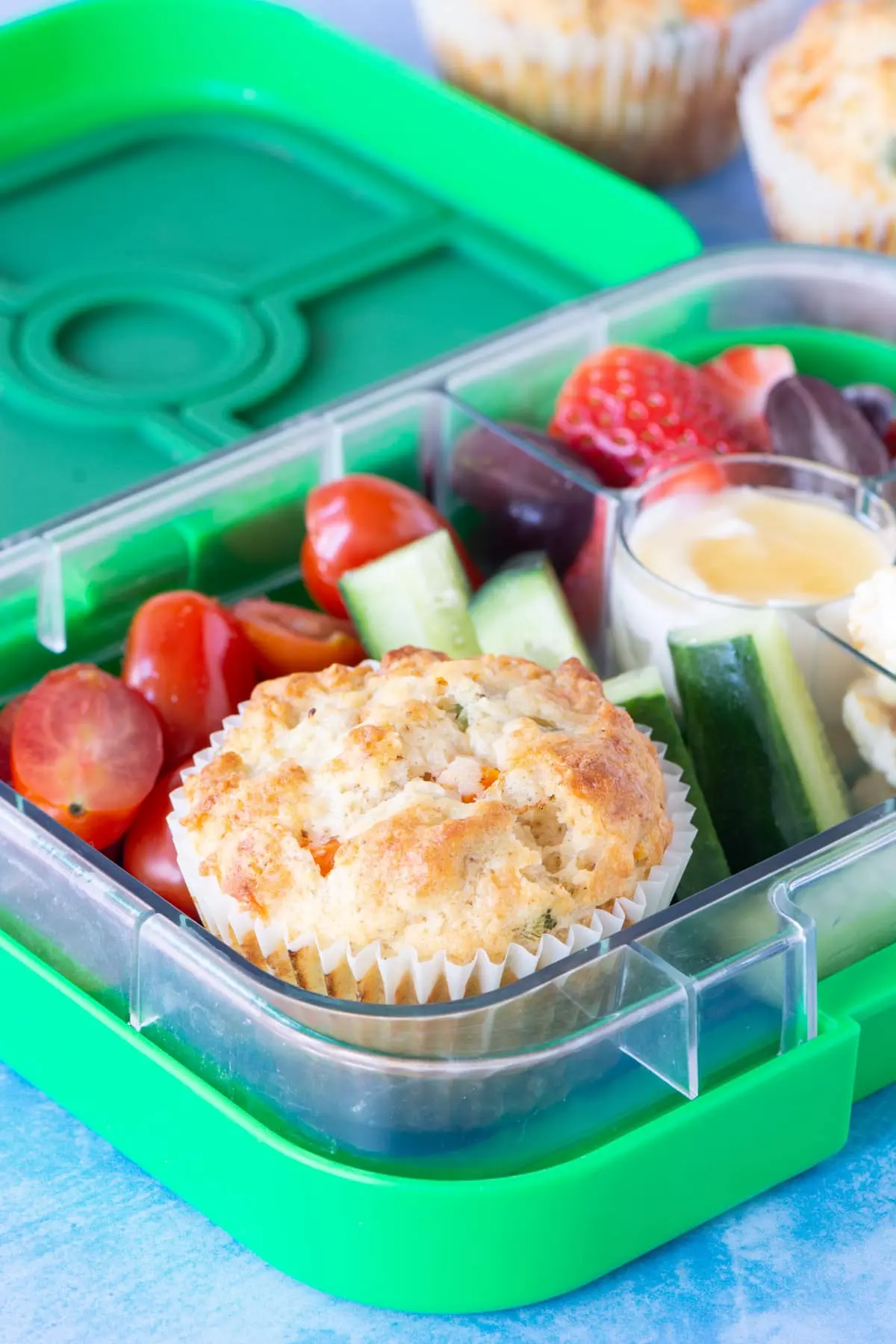 image of a green bento box packed with a mini muffin, cucumber slices, tomatoes, fruit and a dip