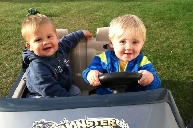 image of two toddler boys driving a motorized toy truck outside.
